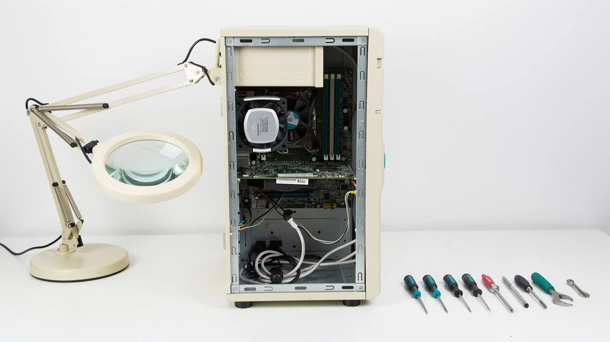 Older desktop computer being serviced on a bench beside a magnifying lamp and a set of precision screwdrivers.
