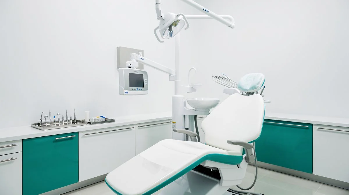 A dental chair with an overhead operating light and a wall-mounted X-ray unit beside a bench of sterile instruments