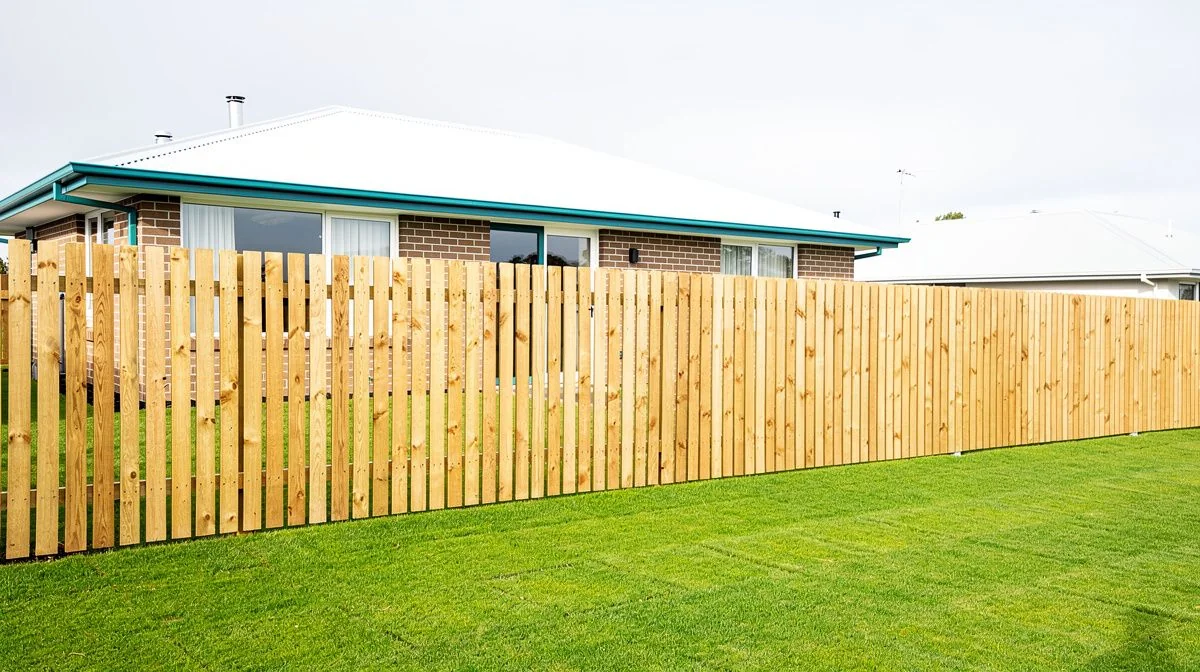 Long straight timber paling fence on flat Christchurch ground with a mown lawn and a single-storey brick house in the background