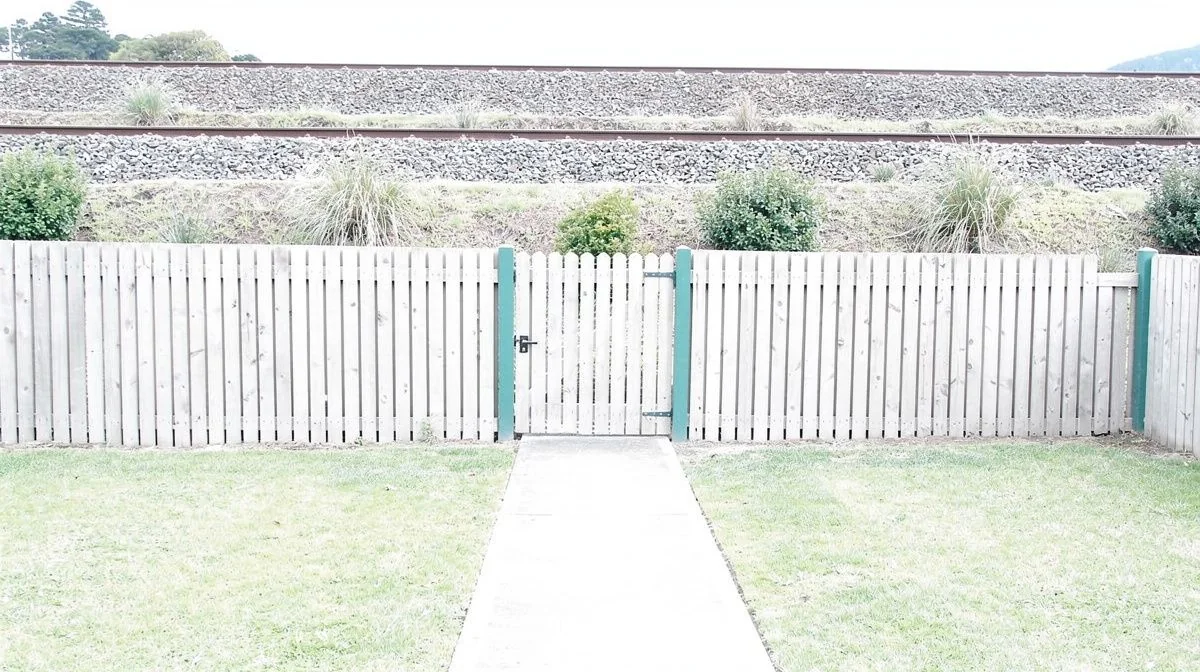 Short timber fence enclosing a compact Upper Hutt backyard with a paling gate, concrete path and railway embankment beyond