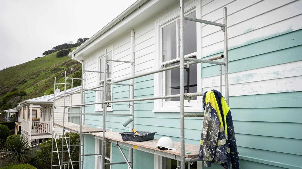 Painter on scaffolding applying a fresh coat to the windward weatherboard wall of a hillside Wellington villa