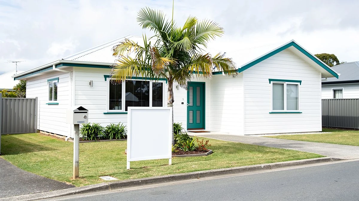 A white weatherboard house on a Tauranga street with a for-sale sign near the letterbox and a mature palm in the front garden