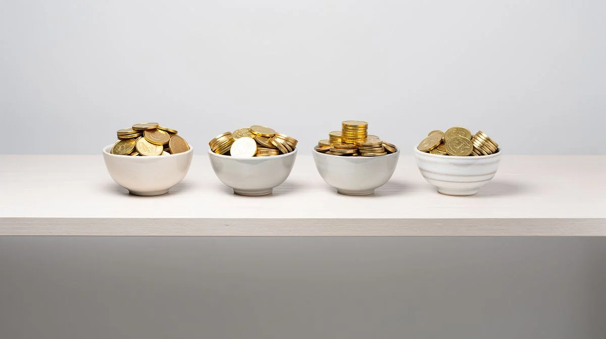 Five small ceramic bowls in a row on a shelf, each filled to a different height with coins