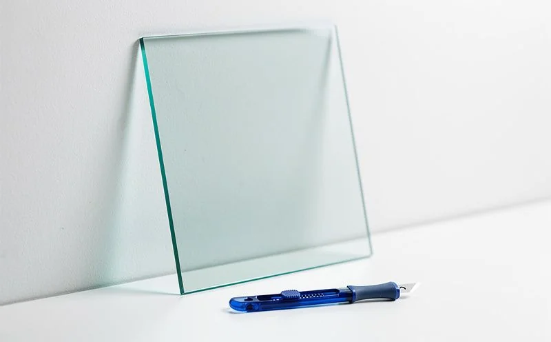 A sheet of clear glass leaning against a pale wall with a blue glass cutter in front