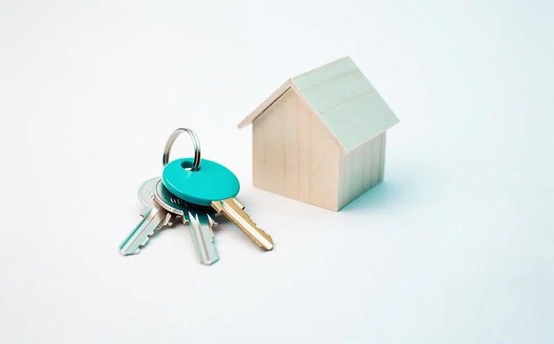 A set of keys on a teal keyring beside a small pale wooden model house